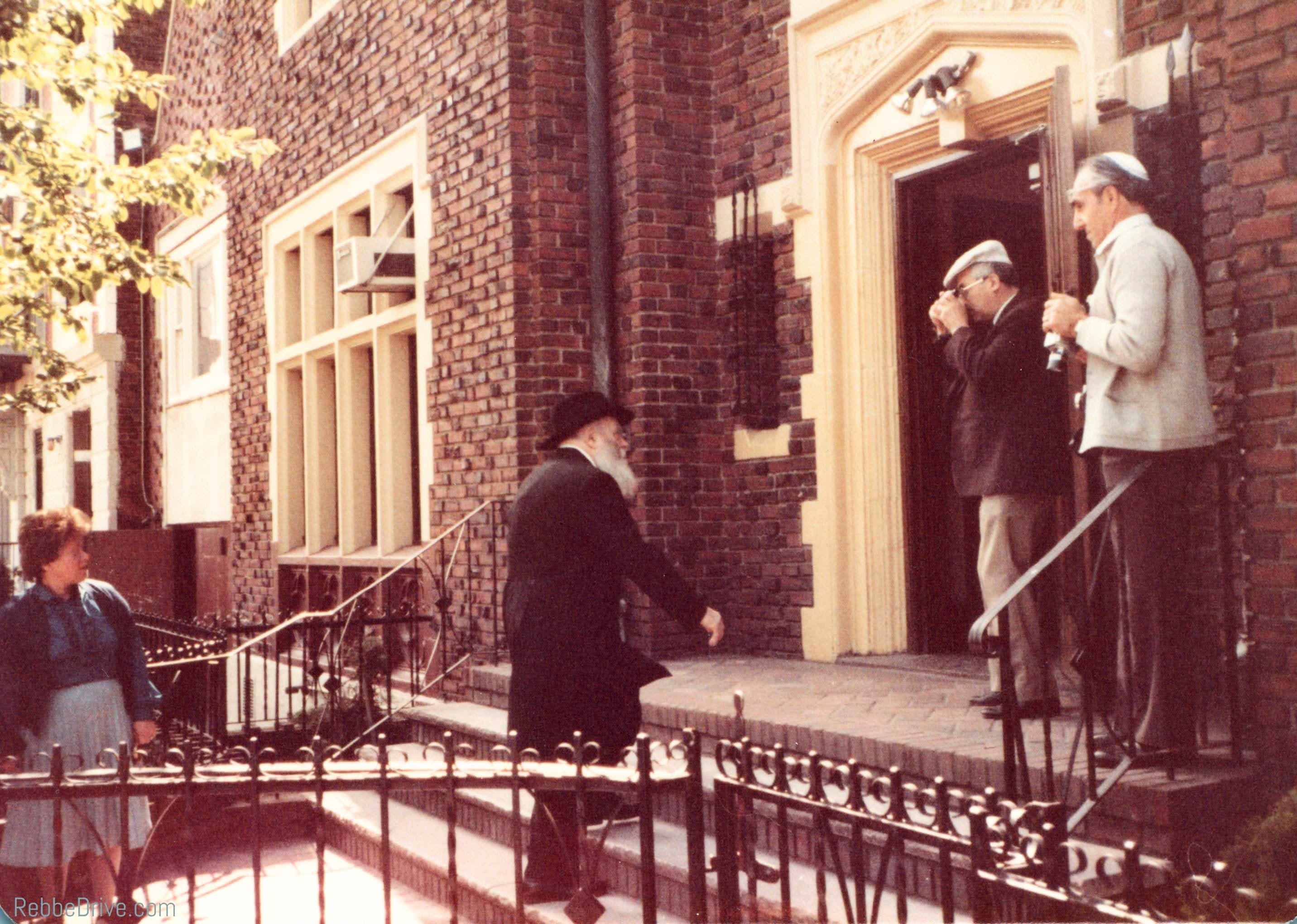 The Lubavitcher Rebbe walking toward the entrance of 770 Eastern Parkway, with people waiting nearby.