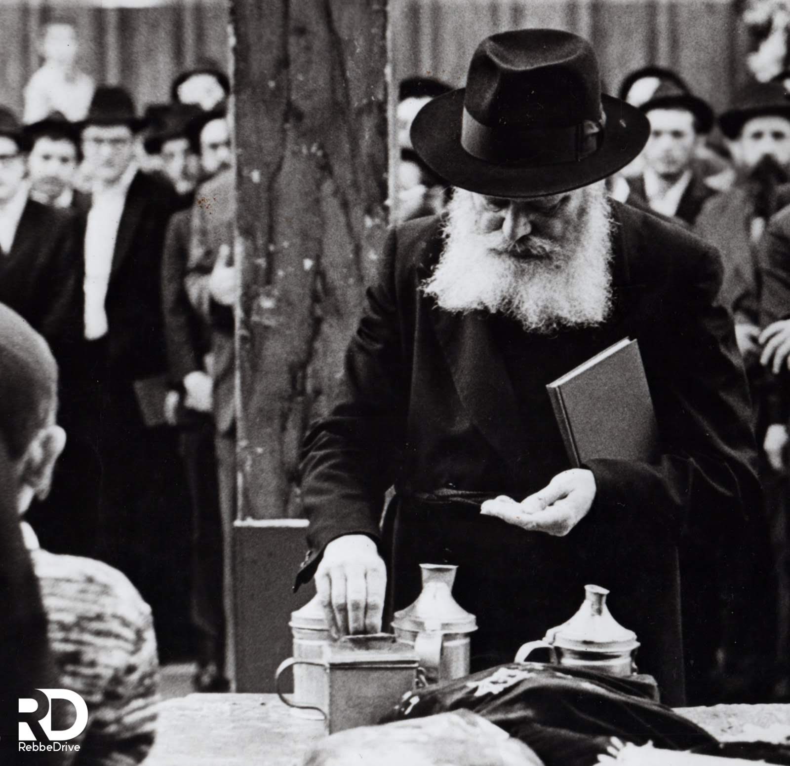 The Lubavitcher Rebbe placing coins into a charity box, holding a book as people gather nearby.